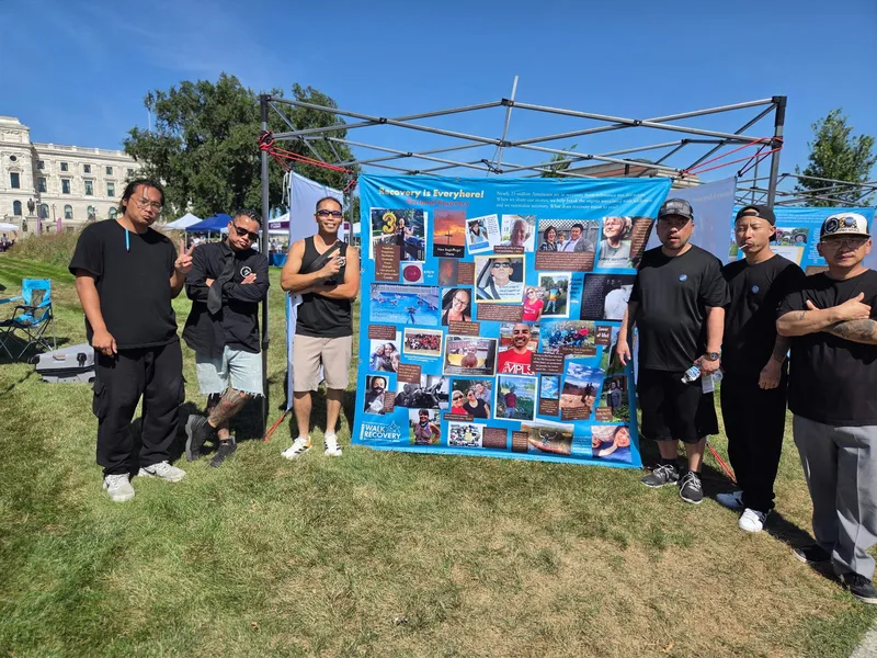 A group standing outdoors beside a large "Recovery Is Everywhere" story banner at an outdoor event